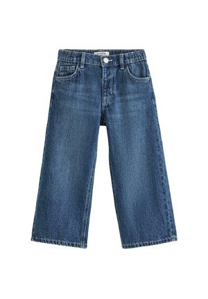 Smilende lille barn med krøllet hår står iført en enkel hvid t-shirt, løse blå jeans og hvide sneakers foran en ensfarvet baggrund.