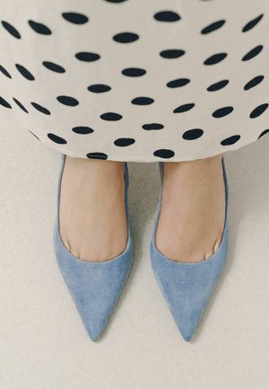 Füße in hellblauen spitz zulaufenden Ballerinas unter einem weißen Rock mit großen schwarzen Punkten auf hellem Boden.