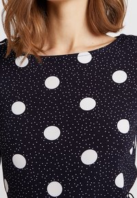 Marinblå tyg toppen med vita prickar och mindre prickmönster. Rundad halsringning, korta ärmar och slät yta.