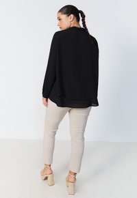 Black long-sleeve blouse with a ruffled neckline, layered design, paired with beige fitted pants and strappy wedge sandals.