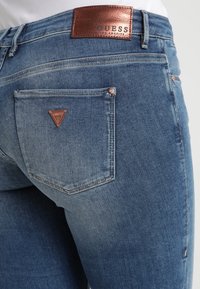 Blå denimjeans med en bakficka som har en kopparfärgad triangul Guess-logotyp och en kopparfärgad Guess Los Angeles-märkning på midjebandet.