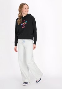Felpa nera con grafica colorata "Spread Love", abbinata a jeans a gamba larga di un blu chiaro e sneakers blu navy. Il tessuto sembra morbido e casual.