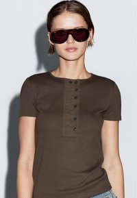 Hnědé žebrované tričko s krátkým rukávem, kulatým výstřihem a knoflíkovou légou. Model nosí oversized tmavé sluneční brýle a pozadí je neutrální.