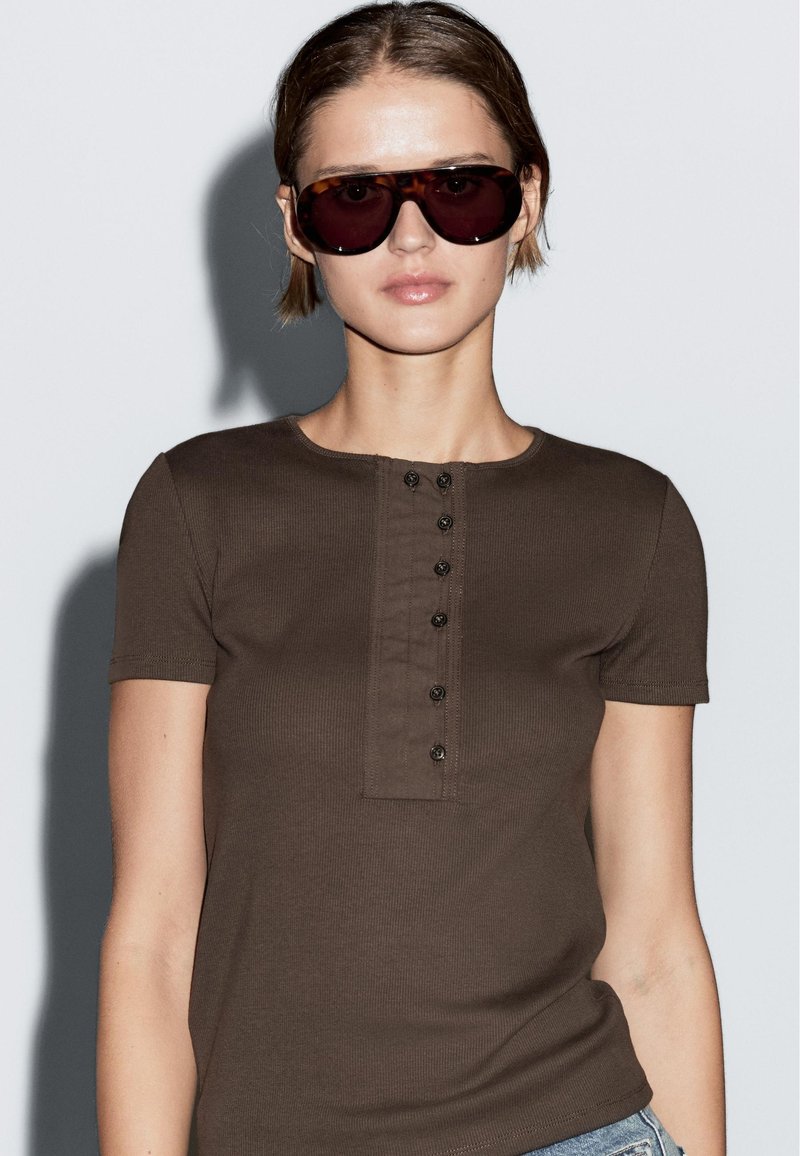 Hnědé žebrované tričko s krátkým rukávem, kulatým výstřihem a knoflíkovou légou. Model nosí oversized tmavé sluneční brýle a pozadí je neutrální.