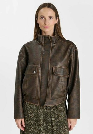 Frau mit langen braunen Haaren, die eine braune Lederjacke und einen Rock mit Leopardenmuster trägt, steht vor einem einfarbigen Hintergrund.