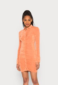 Vestido de camisa de veludo em pêssego, com botões na frente, mangas longas e detalhes franzidos nas laterais para uma silhueta ajustada.