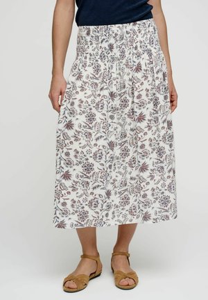 Vrouw met een witte midi-rok met paarse bloemenprint, een gerimpelde tailleband en platte sandalen met een beige gevlochten band.