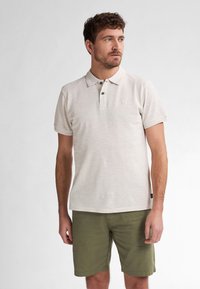 Ljust beige polo med klassisk krage, tvåknapps-knäppning och liten logotyp, matchat med olivgröna shorts. Mjuk, strukturerad tyg.
