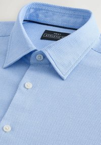 Hellblaues Sportshirt aus strukturiertem Stoff, mit einem Button-Down-Kragen und zwei weißen Knöpfen am Hals. Das Etikett trägt die Aufschrift "NEXT."