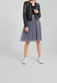 Giubbotto in pelle nera da motociclista con borchie argentate, abbinato a una gonna midi in tulle grigia con un motivo a pois e sneakers bianche.
