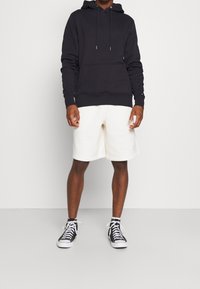 Schwarzer Kapuzenpullover mit Kordelzug, kombiniert mit cremefarbenen Shorts. Die Schuhwerk besteht aus schwarz-weißen High-Top-Sneakern mit Schnürsenkeln.