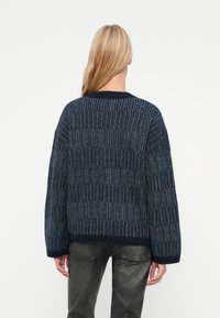 Femeie cu păr lung blond, purtând un pulover tricotat texturat de culoare albastru închis și pantaloni negri estompați, întorcându-se cu spatele, pe un fundal alb simplu.