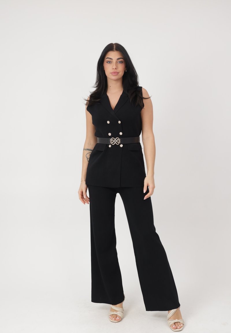 Mujer con cabello largo y negro que lleva un blazer negro sin mangas con botones decorativos, pantalones anchos negros y sandalias beige, de pie frente a un fondo blanco.