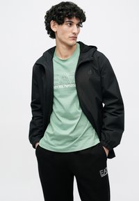 Fekete kapucnis dzseki, mentazöld pólóval, amelyen fehér grafikák vannak, fekete nadrággal, melyen logó található. Simább anyag, laza szabás.