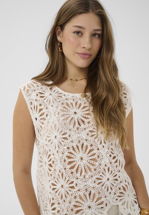 Mladá žena s dlhými vlasmi, ktorá má na sebe biely háčkovaný kvetinový top bez rukávov a zlaté šperky, pózujúca pred jednoduchým pozadím.