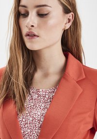 Oranžový blazer s klopami, cez bielu blúzku s kvetinovým vzorom. Štruktúrovaná tkanina pridáva hĺbku, na uchu sú viditeľné zlaté náušnice.