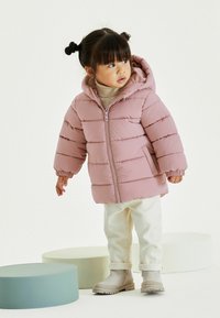 Doudoune rose clair avec capuche, fermeture éclair et poignets côtelés. Associée à un pantalon crème et des bottes claires, se tenant sur des accessoires circulaires doux.