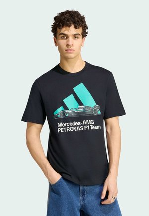 Młody mężczyzna z kręconymi włosami, w czarnej koszulce Mercedes-AMG Petronas F1 Team i niebieskich dżinsach, stoi z jedną ręką w kieszeni.