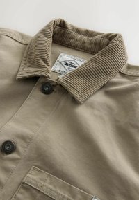 Beige corduroy collar shirt with button closure, featuring a woven label and a textured finish. Includes front pocket detail.