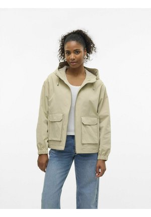 Veste beige à capuche en tissu lisse, dotée de poignets élastiques, de deux poches avant et d'une fermeture éclair à l'avant. Portée par-dessus un haut blanc.