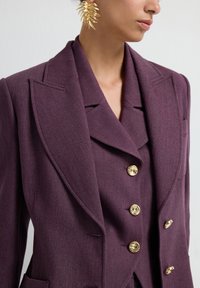 Paarse dubbele blazer met opvallende revers, gouden knopen en een gestructureerde afwerking. Opvallende oorbel in de vorm van een blad.