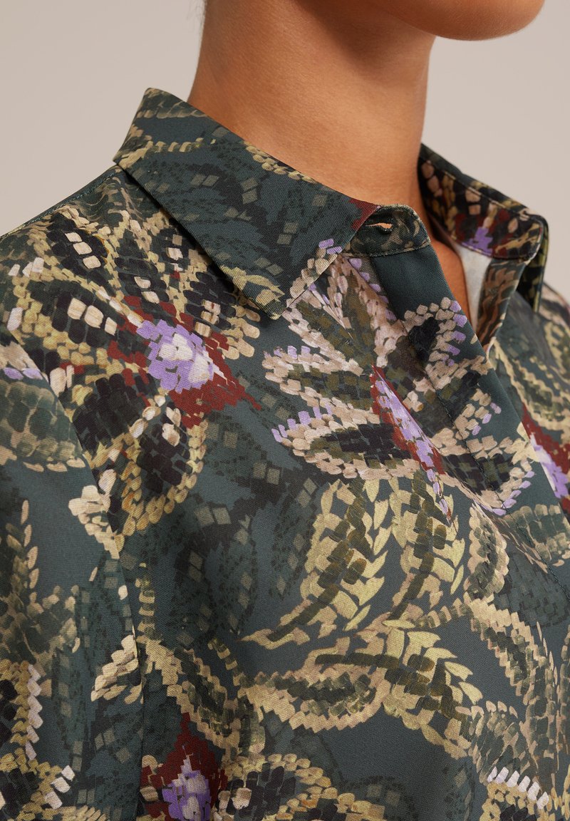 Een blouse met kraag in een donkere groene stof met een bloemenpatroon in tinten paars, rood en goud, met een gladde textuur.