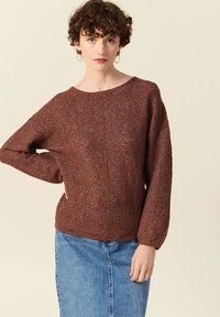 Brauner, strukturierter Strickpullover mit weitem Ausschnitt und Ballonärmeln, kombiniert mit einem Jeansrock. Beide Kleidungsstücke haben lässige Designs.