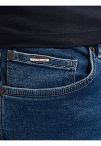 Vaqueros de mezclilla azul con un logo de PETROLIND en el bolsillo para monedas, con costuras en contraste y una textura ligeramente desgastada.