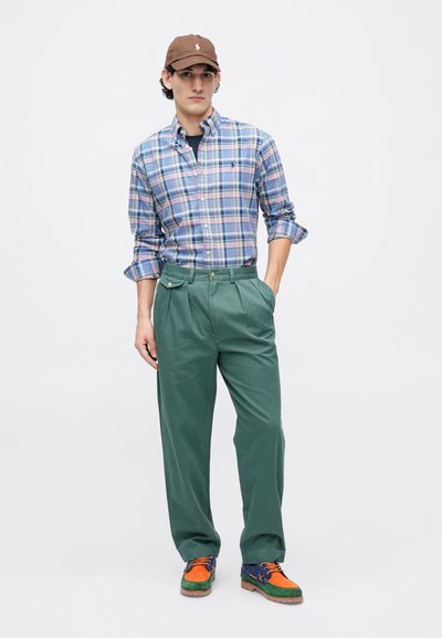 Καρό πουκάμισο σε μπλε και πράσινο με μακριά μανίκια, πράσινο πλισέ παντελόνι και πολύχρωπα παπούτσια με πορτοκαλί και μπλε λεπτομέρειες. Καφέ καπέλο.