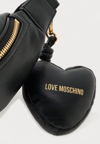 Juodas odinis kryžminis krepšys su auksine užtrauktuko detalė ir širdelės formos pakabuku. Pakabuke rašoma "LOVE MOSCHINO" auksine spalva ant lygaus paviršiaus.