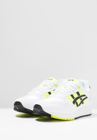 Scarpe da ginnastica bianche con accenti neri e giallo neon, caratterizzate da un design con lacci e suola testurizzata per una maggiore aderenza.