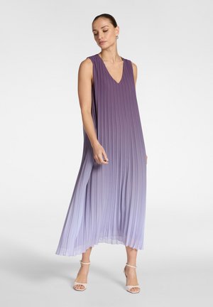 Donna che indossa un abito midi senza maniche, plissettato, con sfumature dal viola al lilla, scollo a V e tacchi bianchi con cinturino alla caviglia, in piedi davanti a uno sfondo semplice.