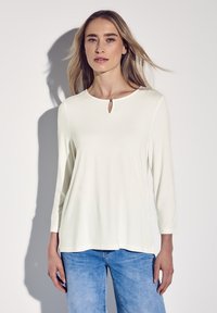 Parte superior de manga larga color crema con escote redondo y un detalle de ojo de cerradura en el centro, combinada con jeans de mezclilla azul. Textura suave y corte relajado.