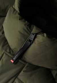 Chaqueta acolchada verde oliva con cuello alto, un tejido exterior texturizado y una correa negra con la marca "SUPERDRY" en el tirador de la cremallera.