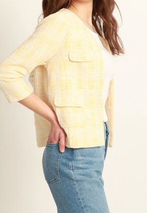 Gelbe karierte Jacke mit Dreiviertelärmeln, strukturiertem Stoff, zwei frontalen Taschen und lockerer Passform, getragen über einem weißen Oberteil und Jeans.