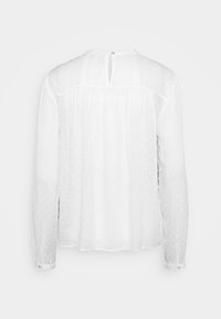 Blouse blanche transparente à manches longues avec texture à pois, empiècement froncé, poignets boutonnés et ouverture goutte avec bouton au dos.