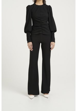 Top nero texturizzato con maniche a palloncino, arricciatura verticale, abbinato a pantaloni neri svasati. Le scarpe bianche con punta a trenino completano il look.