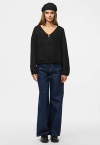 Maglione nero con zip e scollatura a V, due tasche frontali e maniche larghe, abbinato a jeans blu scuro larghi e scarpe nere.