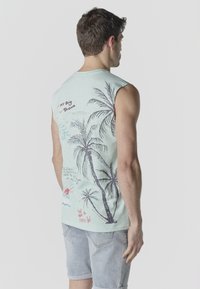 Hombre con camiseta sin mangas azul claro con estampado de palmeras y pantalones cortos de mezclilla desteñidos, de pie de espaldas contra un fondo liso.