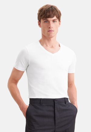 Jeune homme aux cheveux courts et bruns portant un t-shirt blanc à manches courtes avec col en V et un pantalon gris foncé, les mains dans les poches.