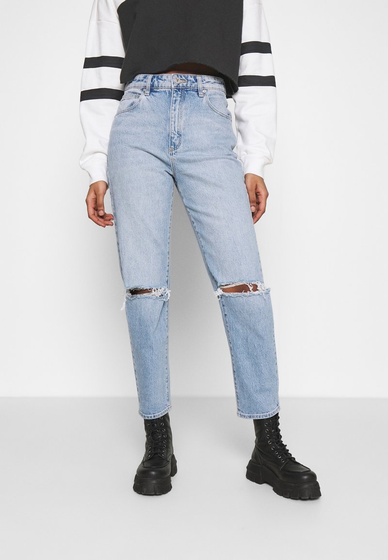 Personne portant un jean bleu clair déchiré aux genoux, des bottes noires épaisses à lacets, et un haut à manches longues noir et blanc avec des rayures sur les manches.