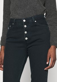 High-waisted black jeans in a denim fabric with five silver buttons and subtle stitching details on the front pockets.