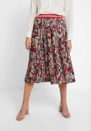 Woman wearing a pleated, floral midi skirt with red and purple tones, paired with a white blouse and beige strappy block-heel sandals.