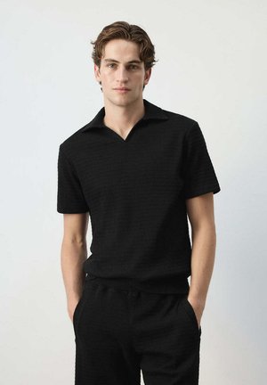 Joven con cabello ondulado lleva una camisa negra de manga corta y pantalones negros a juego con las manos en los bolsillos, de pie frente a un fondo liso.