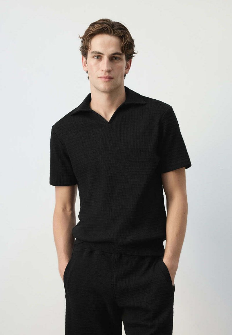Jeune homme aux cheveux ondulés portant une chemise noire à manches courtes et un pantalon noir assorti, les mains dans les poches, debout devant un fond uni.