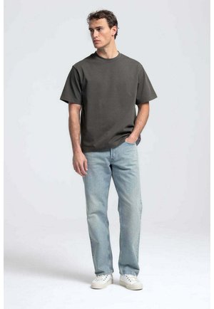 Jonge man die staat met één hand in zijn zak, draagt een donkergrijs oversized T-shirt, lichtblauwe spijkerbroek en witte sneakers op een effen achtergrond.