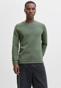 Young man with short hair wearing a green long-sleeve sweater and black jeans, standing against a plain white background.