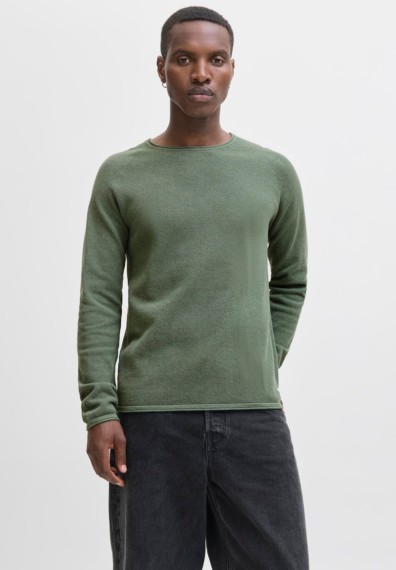 Young man with short hair wearing a green long-sleeve sweater and black jeans, standing against a plain white background.