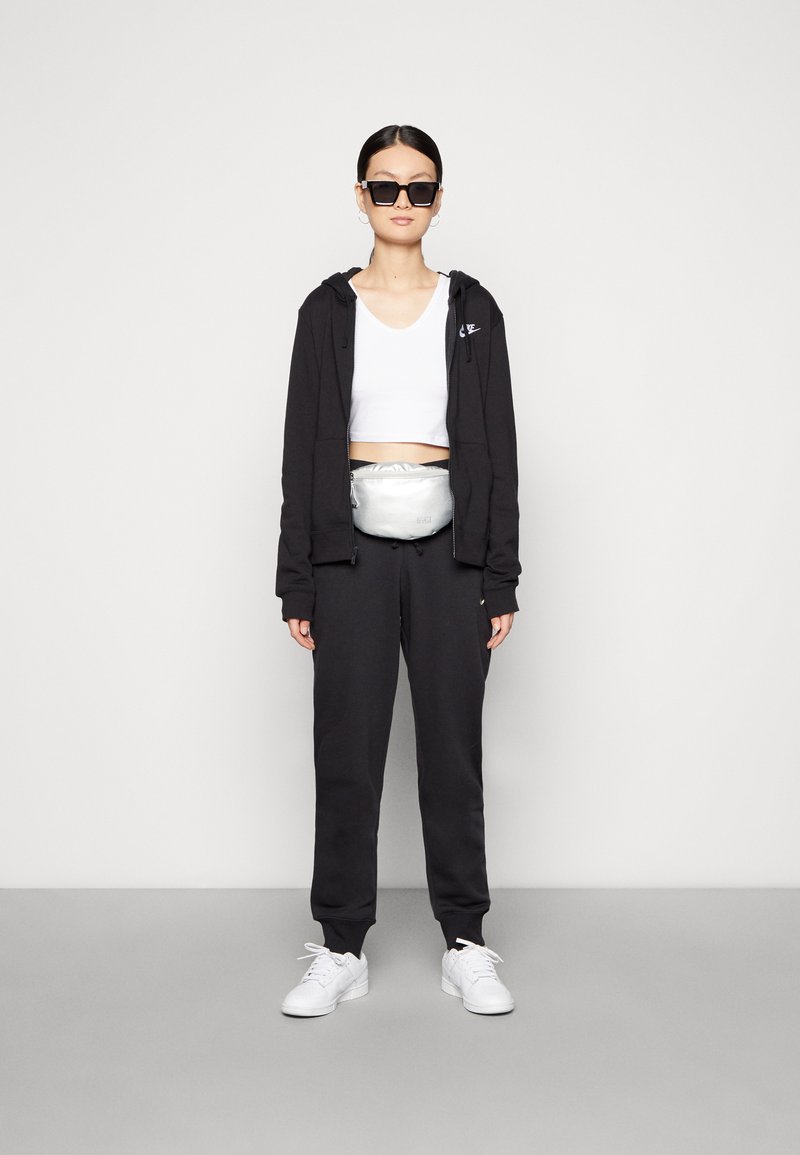 Sudadera con cremallera negra, top corto blanco, pantalones de chándal negros, riñonera plateada, gafas de sol negras de gran tamaño y zapatillas blancas.