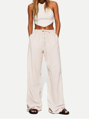 Kvinde iført en hvid tekstureret halter crop top, beige bukser med vid ben og snøre, og sorte sandaler med åben tå, stående med hænderne i lommerne.